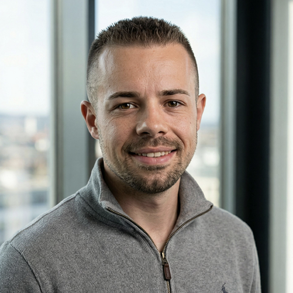 Smiling man with short hair and beard wearing a grey quarter-zip sweater in front of a window.